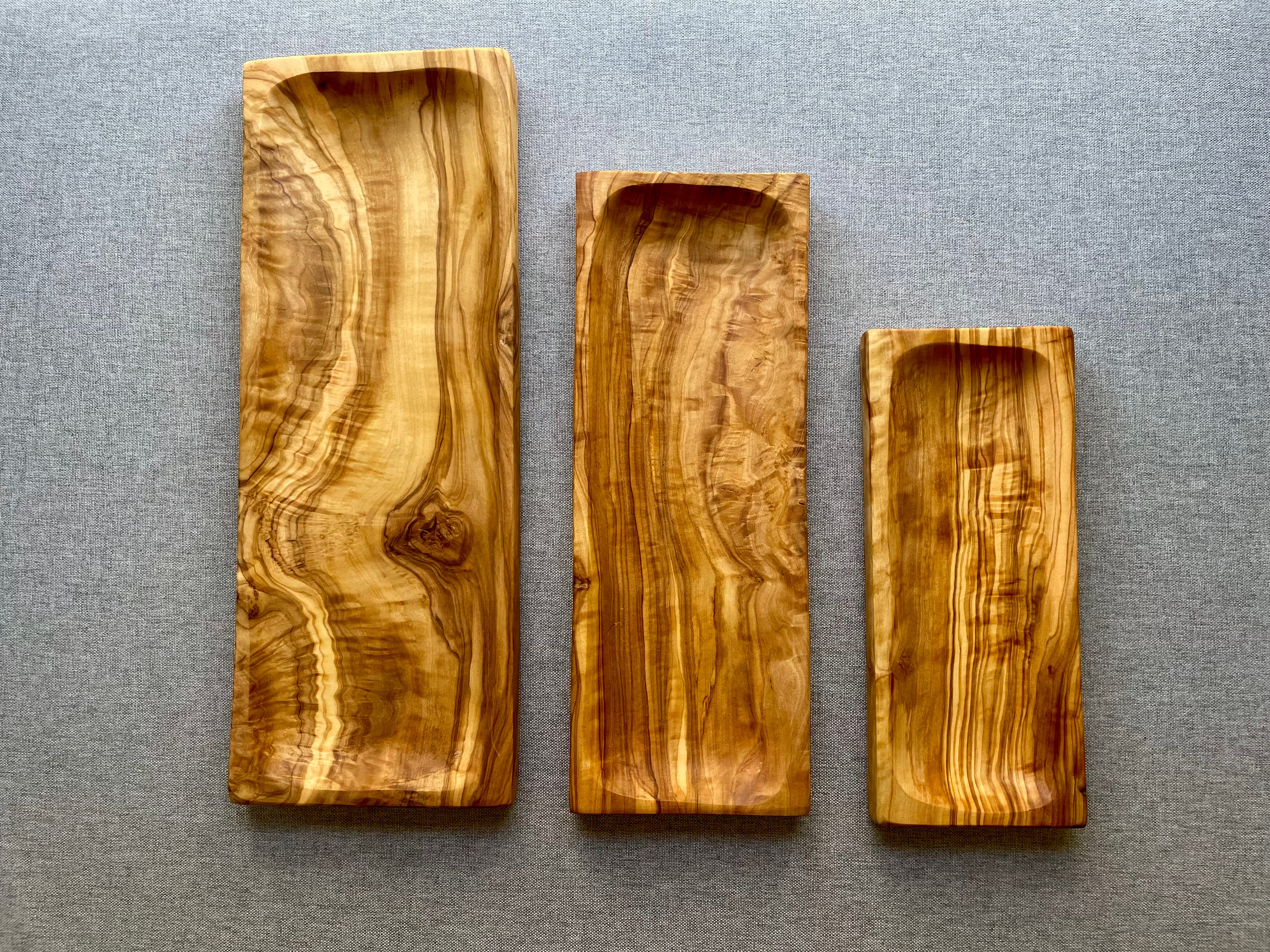 Three olive wood sushi serving trays in small, medium, and large sizes on a gray fabric background - beautifully grained and hand-made from natural olive wood.