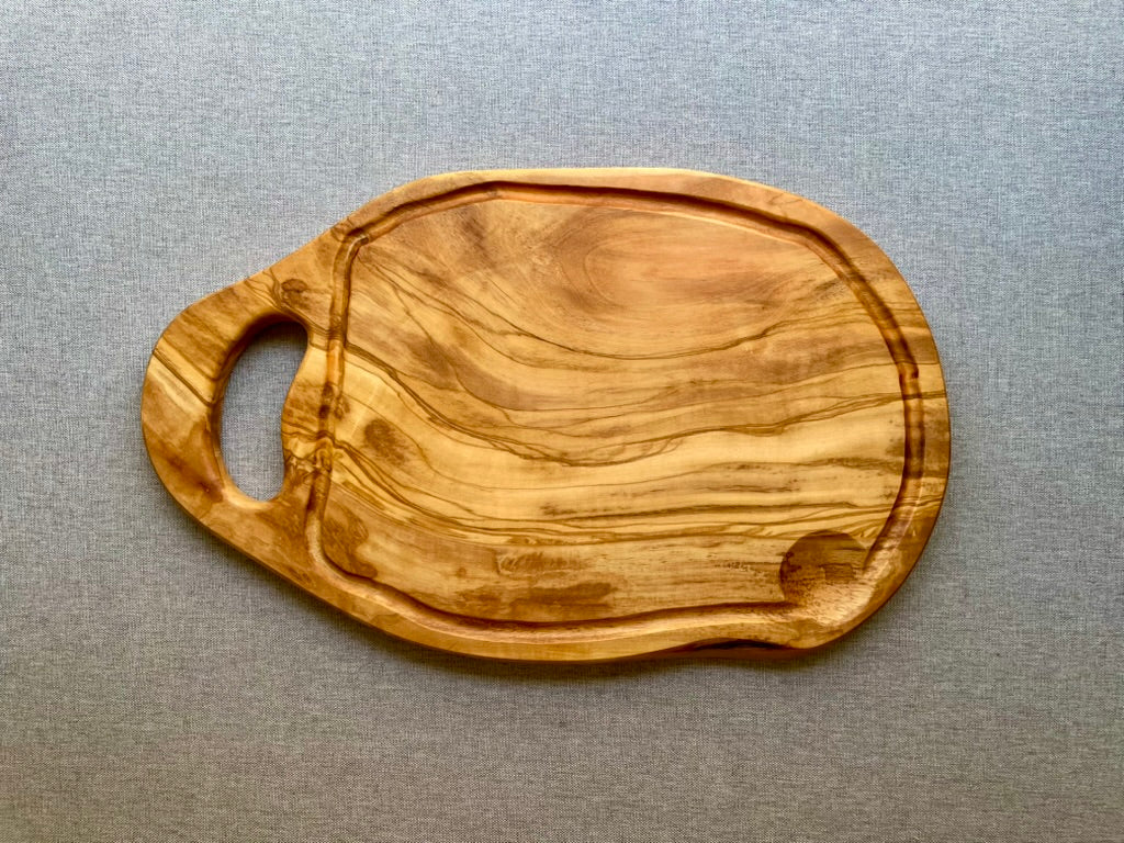 Olive wood cutting and serving board with a carved handle and juice groove on a gray fabric background - hand-made from natural olive wood.