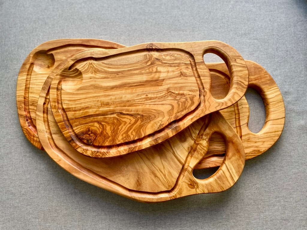 Three nested olive wood cutting and serving boards with carved handles and grooves, stacked on a gray fabric background - hand-made from natural olive wood.