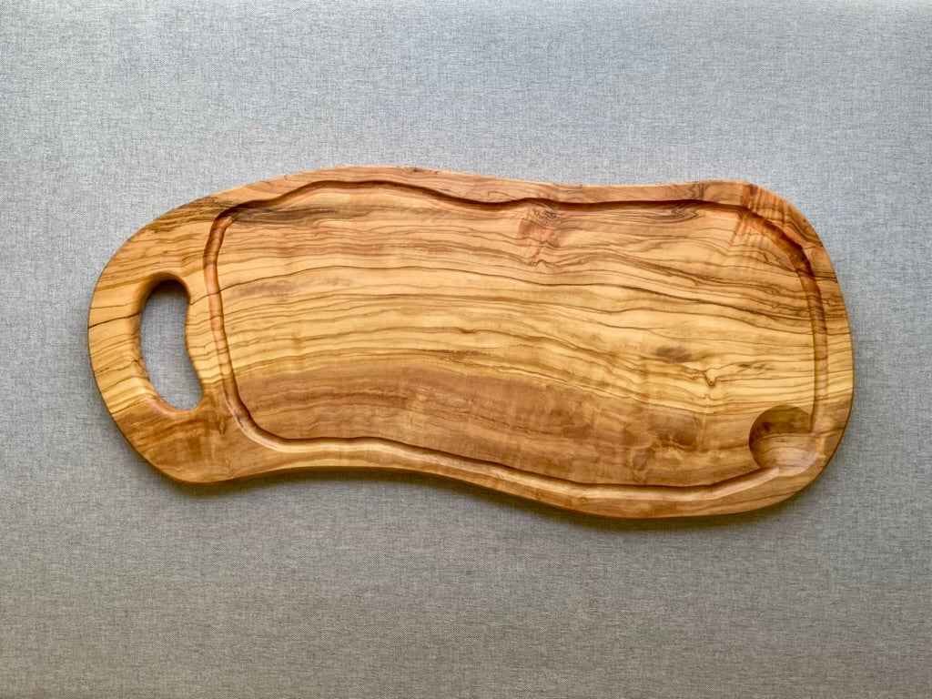 Olive wood cutting and serving board with a natural live edge, carved handle, and juice groove on a gray fabric background - hand-made from natural olive wood.