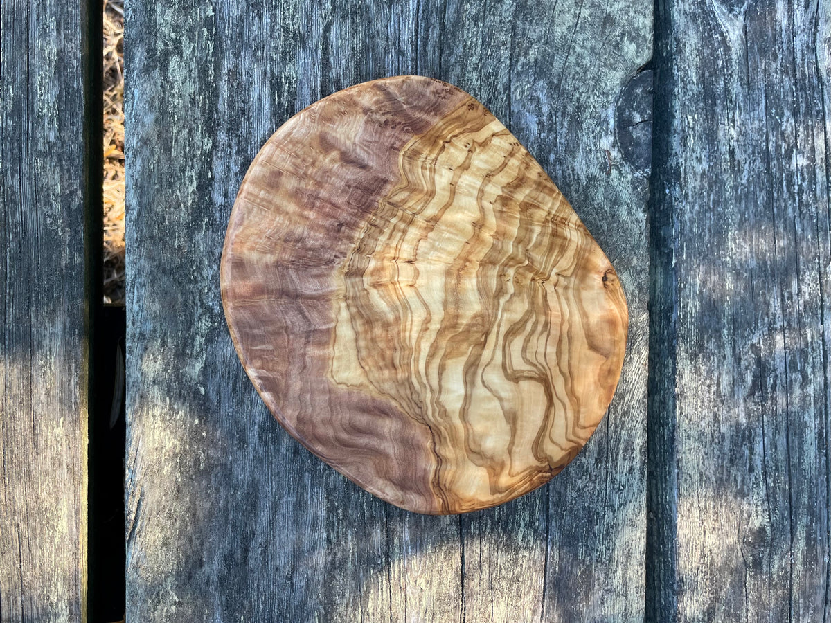 A round olive wood trivet with wavy grain on weathered blue-gray wood planks - hand-made from natural olive wood.