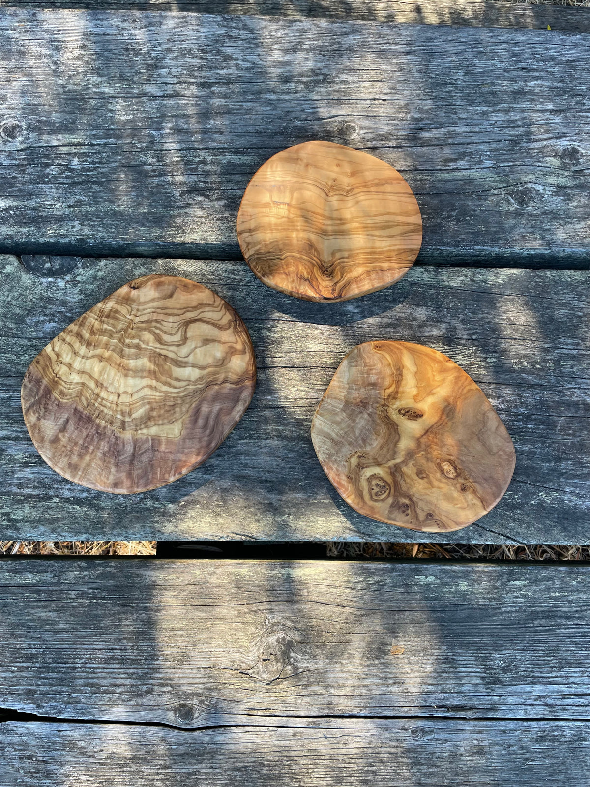 Three round olive wood trivets with swirling grain on weathered gray wood planks - hand-made from natural olive wood, each one unique.