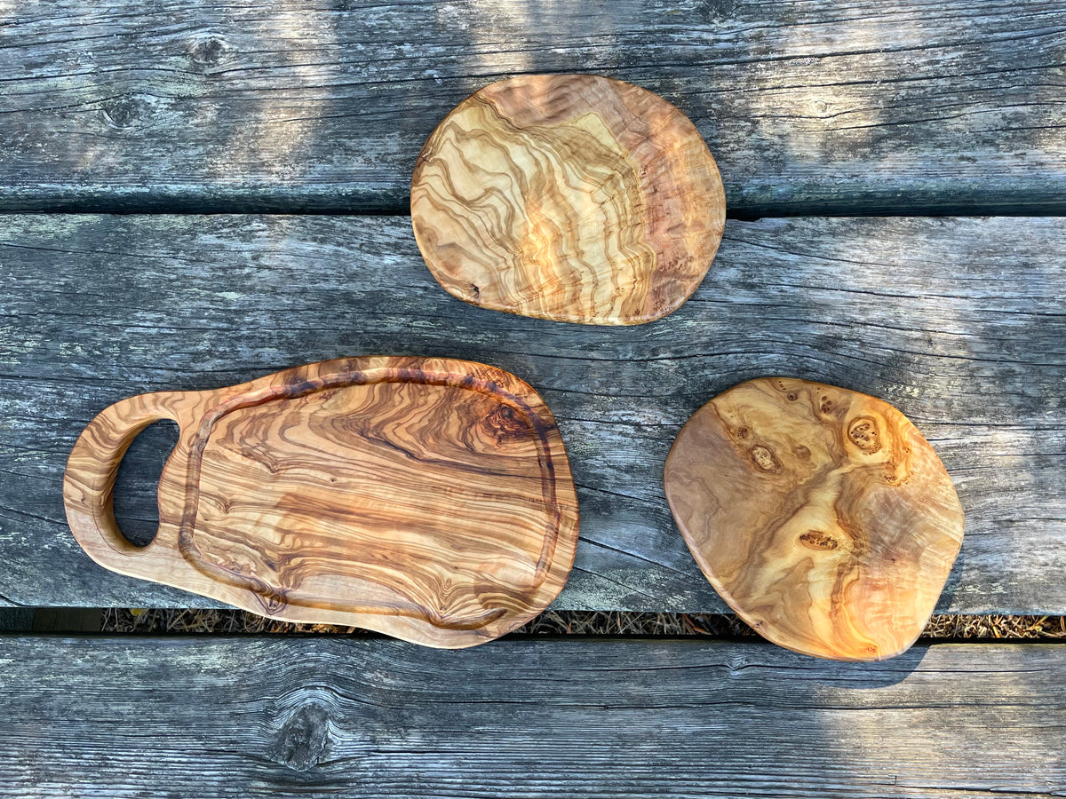 A round olive wood trivet, an olive wood cutting board with handle and groove, and an olive wood serving board arranged on a weathered outdoor table - all hand-carved from natural olive wood.