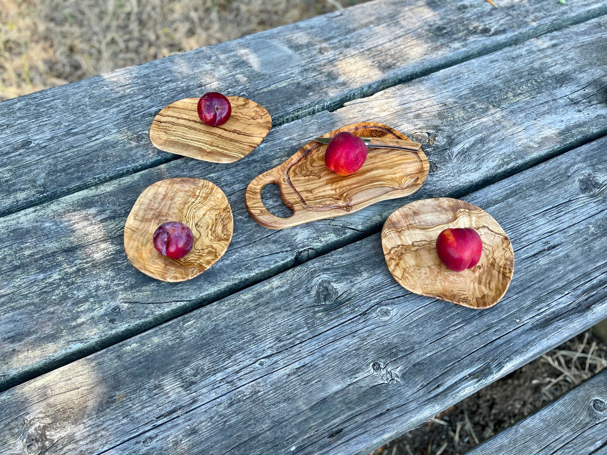 Four olive wood serving pieces - a cutting board with handle and three thin oval plates - each styled with a red plum on a weathered outdoor table, all hand-carved from natural olive wood.