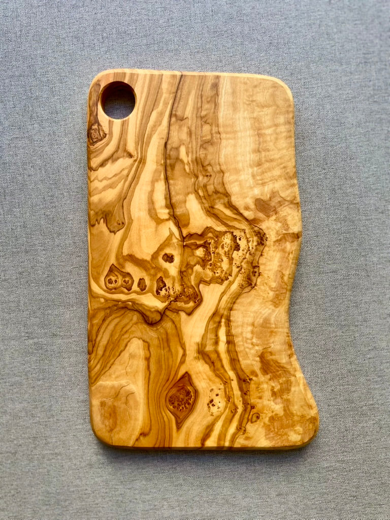 A rectangular olive wood cutting and serving board with striking wavy grain and a hanging hole, shown on a gray surface - hand-made from natural olive wood, perfect for preparing and serving snacks.