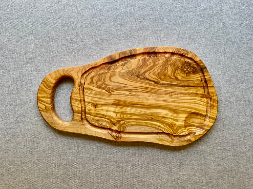 Olive wood cutting and serving board with a carved handle and juice groove, shown on a gray fabric background - hand-made from natural olive wood with rich, one-of-a-kind grain.