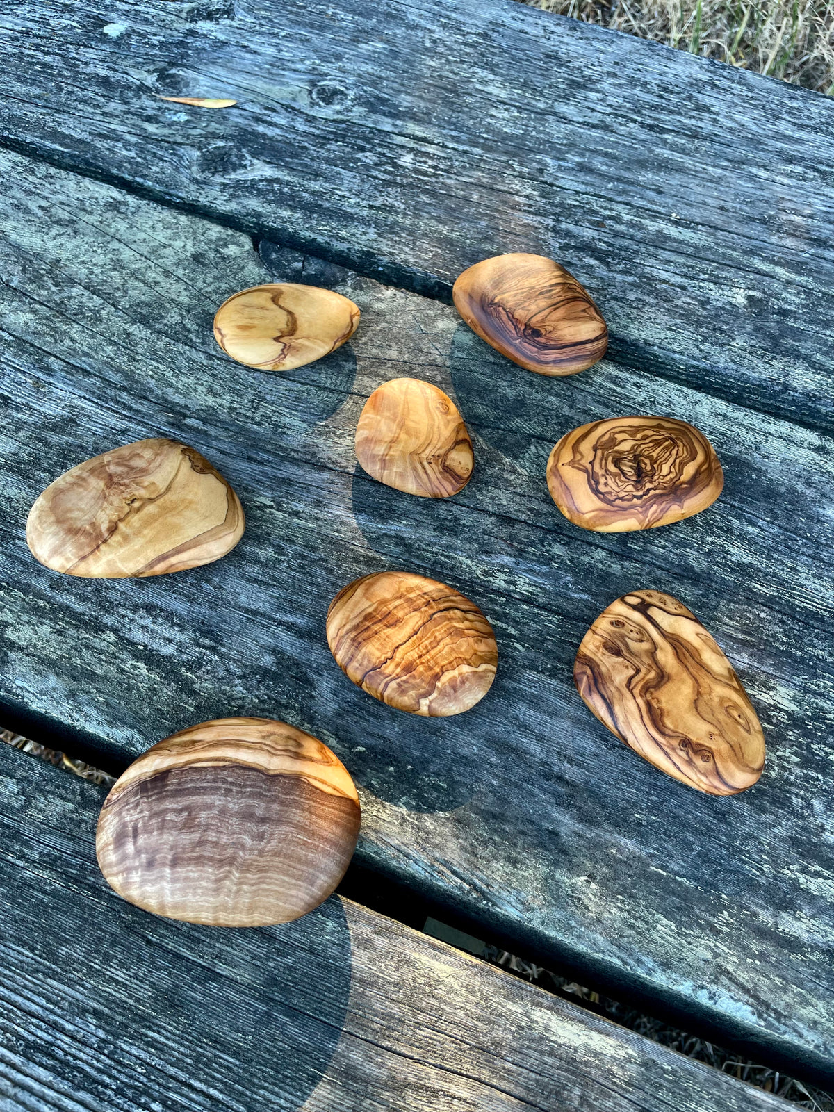 A set of eight smooth olive wood cairn stacking stones laid out on a weathered wood table, each hand-cut from natural olive wood with unique grain - a decorative gift and toy for all ages.
