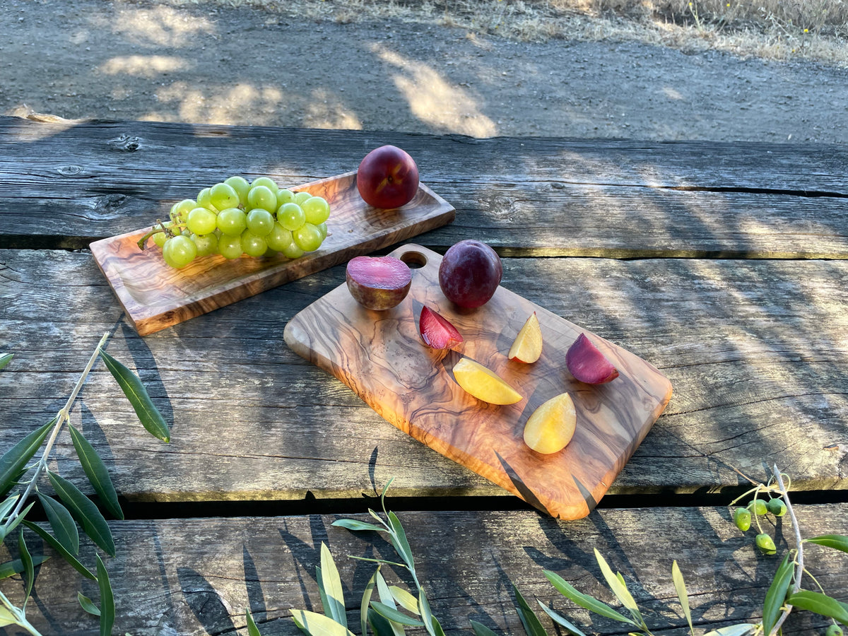 Two olive wood serving boards styled with fresh fruit on a weathered outdoor table - a rectangular olive wood board with sliced plums and peaches, and a longer olive wood serving tray holding green grapes. Both hand-carved from natural olive wood - pair them to set a beautiful table.