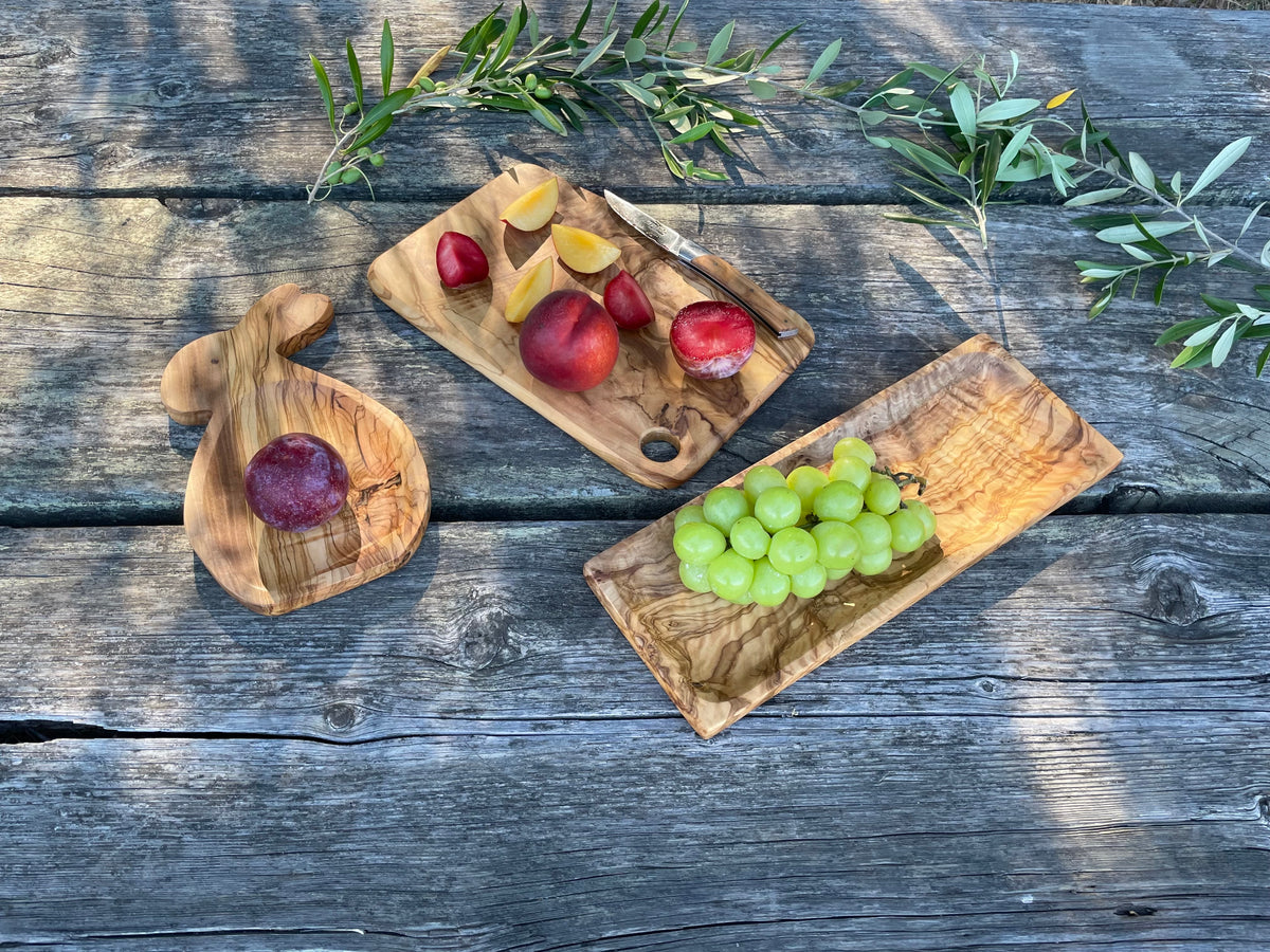 Three handcrafted olive wood pieces styled with fresh fruit on a weathered picnic table: our olive wood sushi tray (right, with green grapes), a rectangular olive wood serving board (center, with sliced peaches and plums), and an olive wood bunny plate (left). Each is hand-carved from natural golden olive wood - mix and match to set a beautiful table.