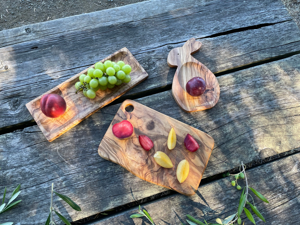 Three handcrafted olive wood pieces displayed with fresh fruit on a weathered picnic table: an olive wood bunny plate (top right, with a plum), a rectangular olive wood serving board (left, with green grapes and a nectarine), and an olive wood cutting board (center, with sliced plums and nectarines). Each is hand-carved from natural olive wood - mix and match to set a beautiful table.