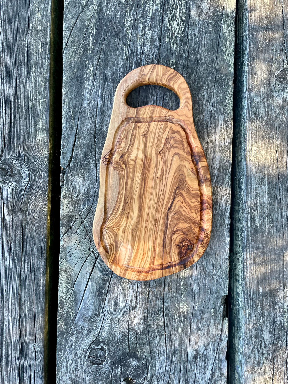 An olive wood cutting and serving board with a rounded handle and juice groove on weathered blue-gray wood planks - hand-made from natural olive wood.