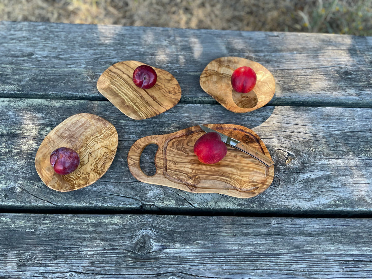 Four olive wood serving pieces - including a cutting board with handle and thin oval plates - styled with red plums and a knife on a weathered outdoor table, all hand-carved from natural olive wood.