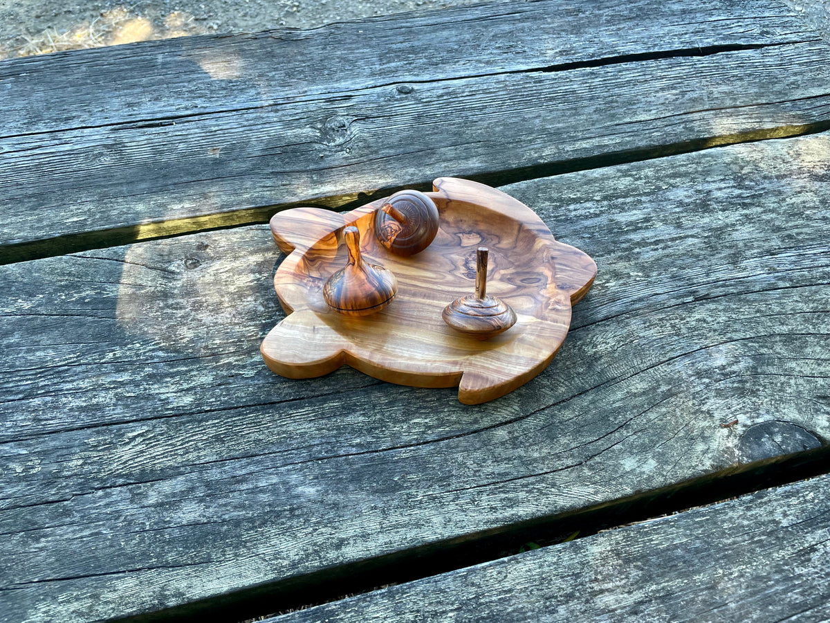 An olive wood turtle-shaped dish holding three olive wood spinning tops on a weathered outdoor table - both hand-carved from natural olive wood.