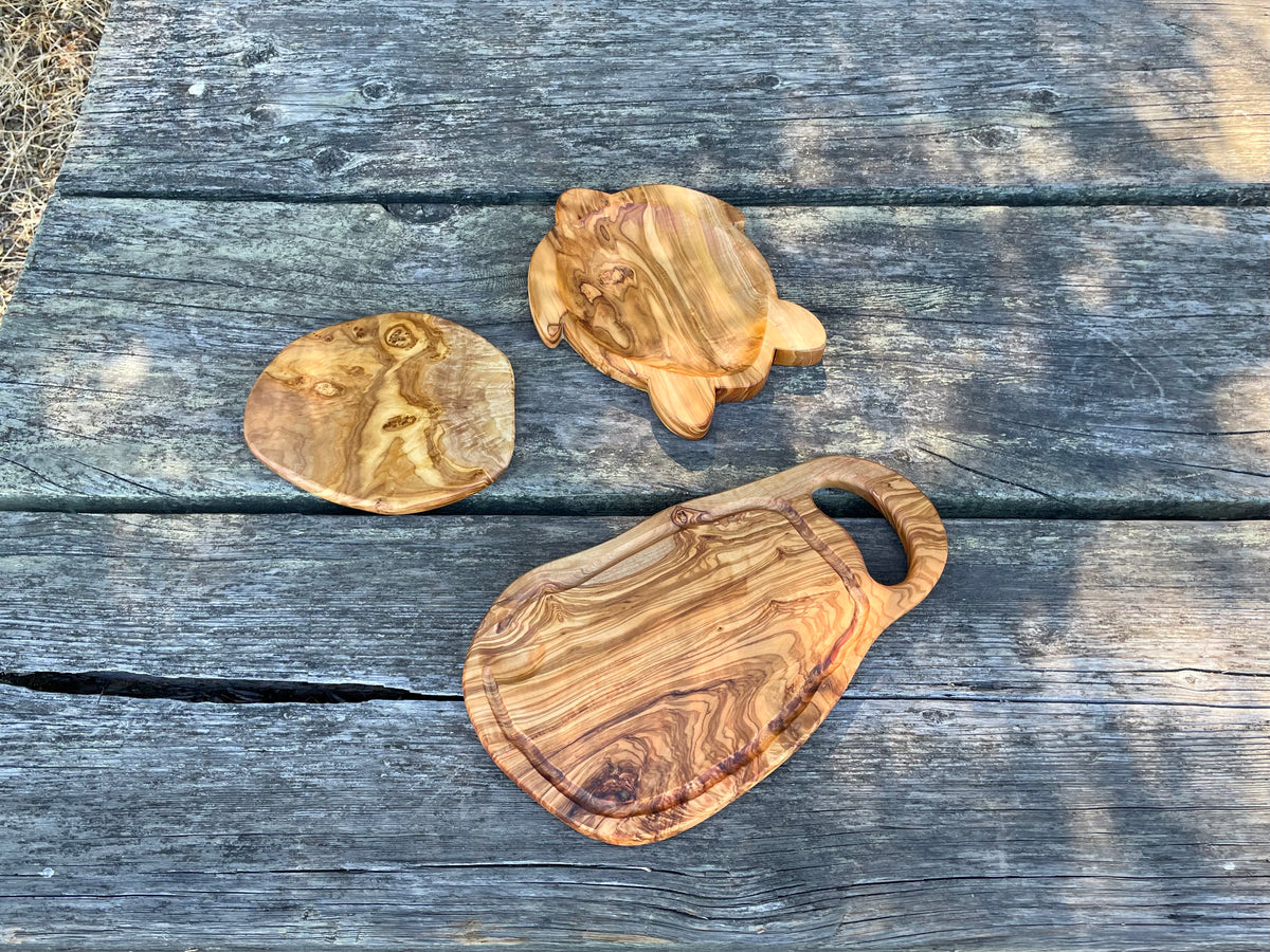 Two olive wood turtle-shaped plates and an olive wood cutting board with handle and groove arranged on a weathered outdoor table - all hand-carved from natural olive wood.