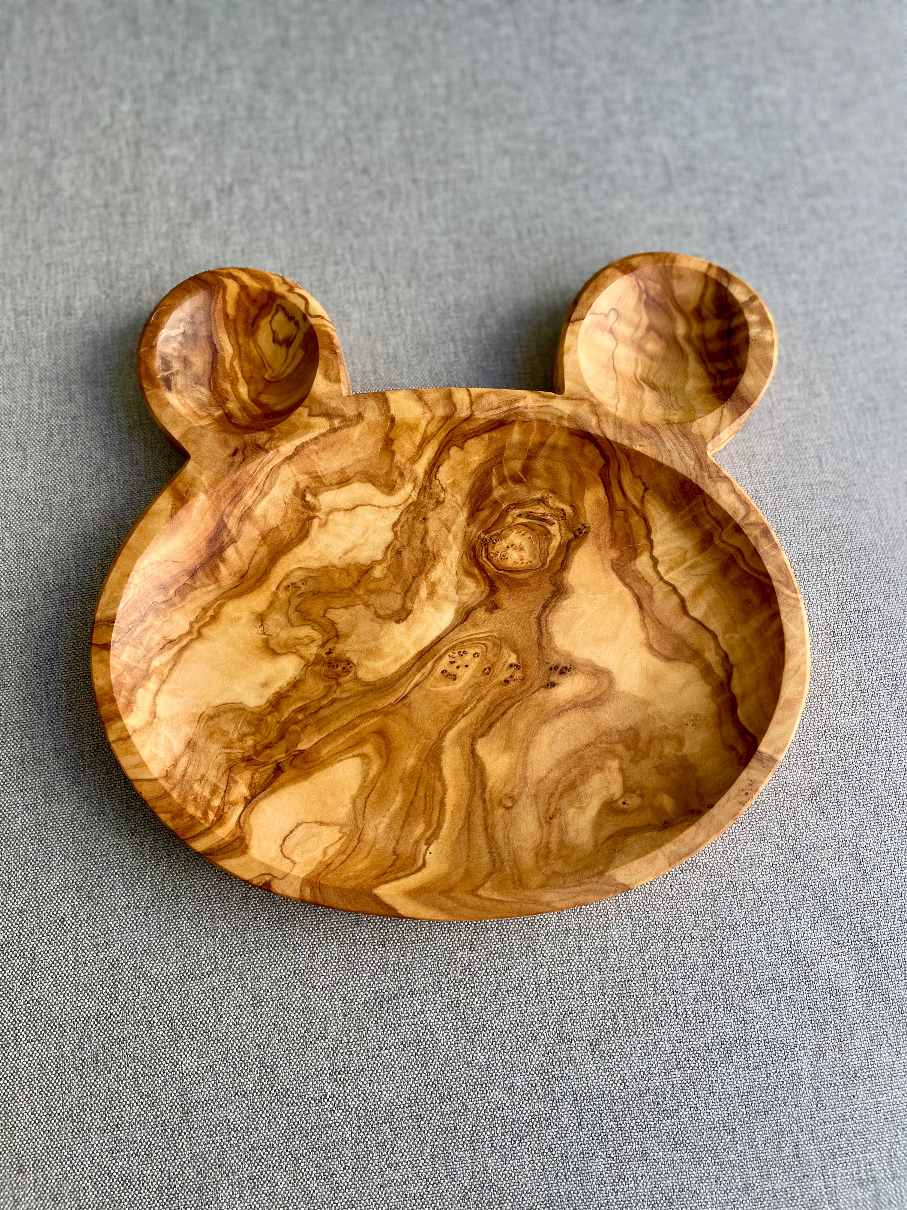 Olive wood bear-shaped plate with a round face and two ears, on a gray fabric background - hand-made from natural olive wood, a fun dish for children's snacks.