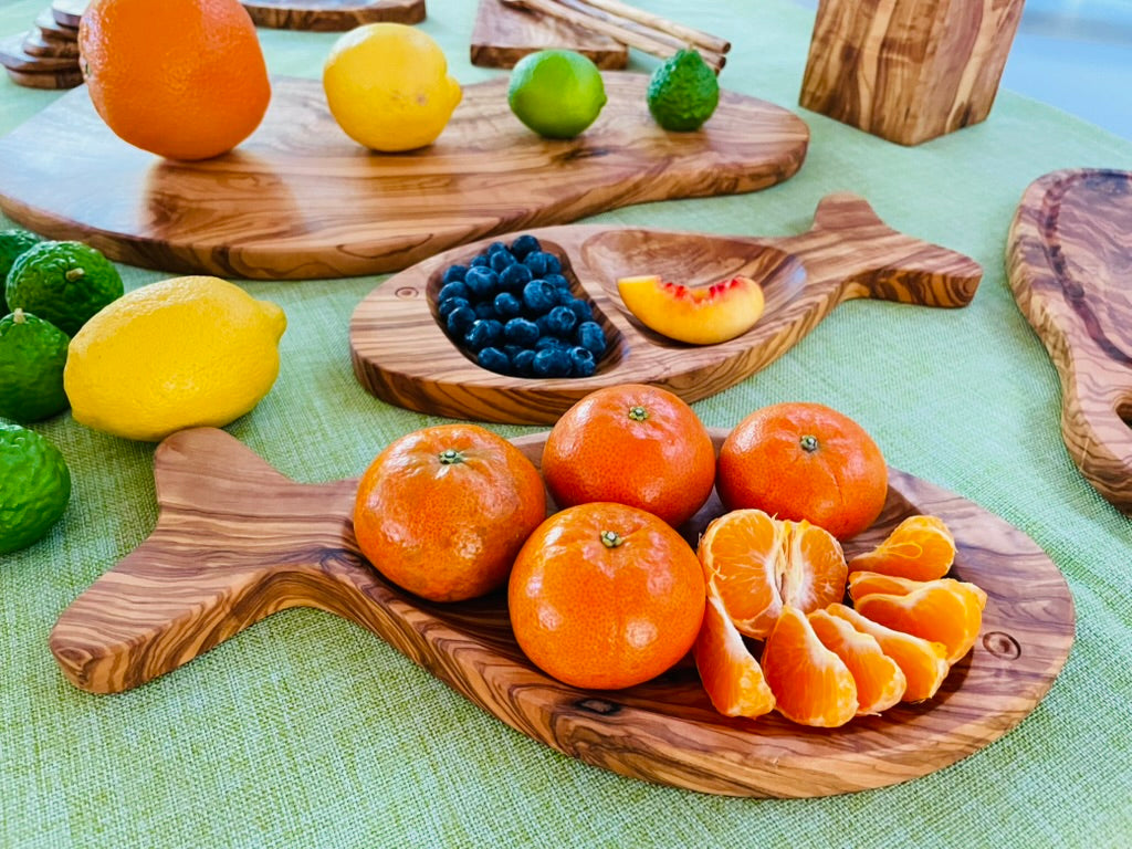 A colorful spread of olive wood fish-shaped serving dishes styled with oranges, lemons, limes, blueberries, and peaches on a green tablecloth, alongside an olive wood serving board - all hand-carved from natural olive wood.
