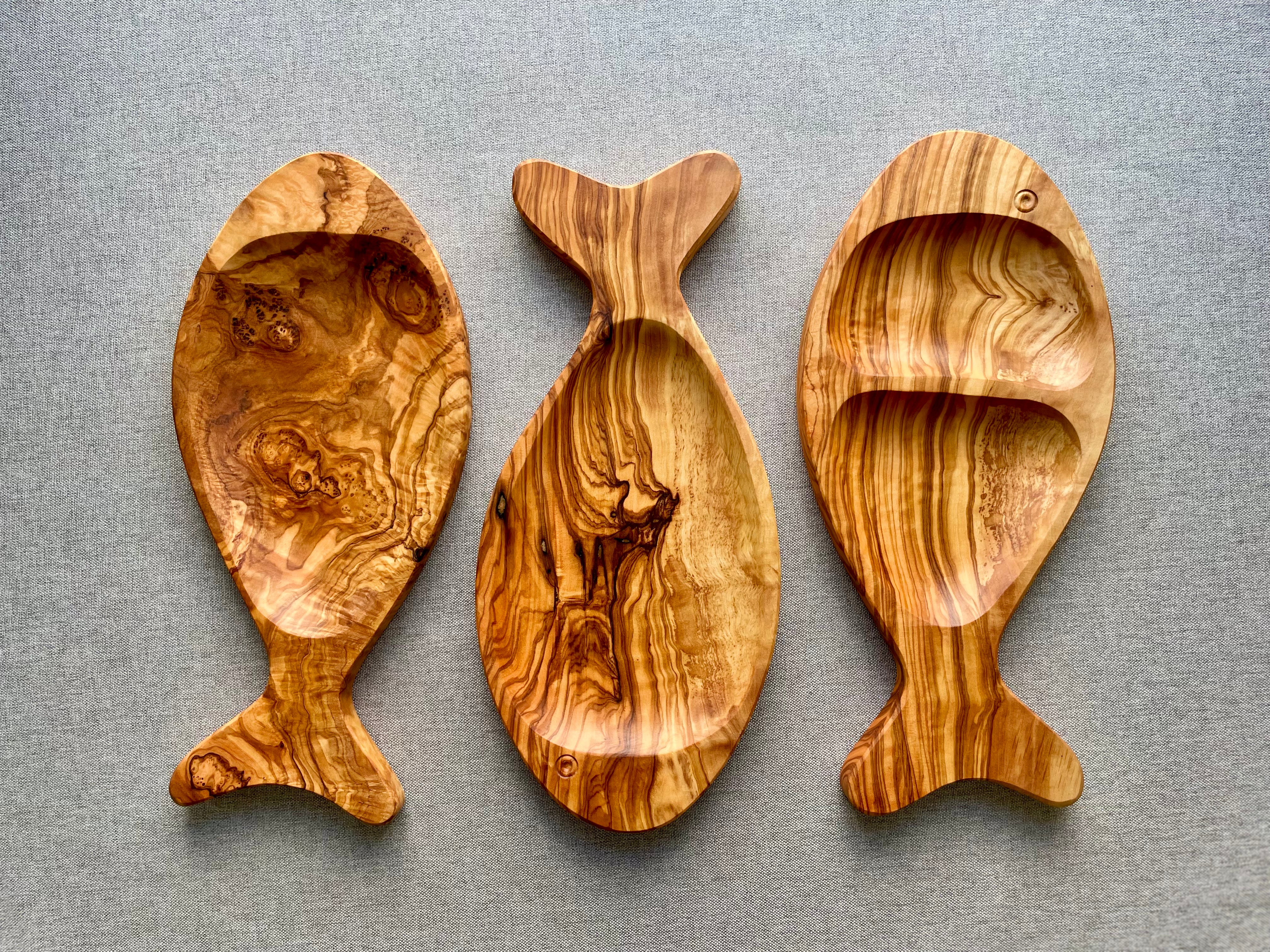 Three olive wood fish-shaped platters in different styles on a gray fabric background - hand-carved from natural olive wood.