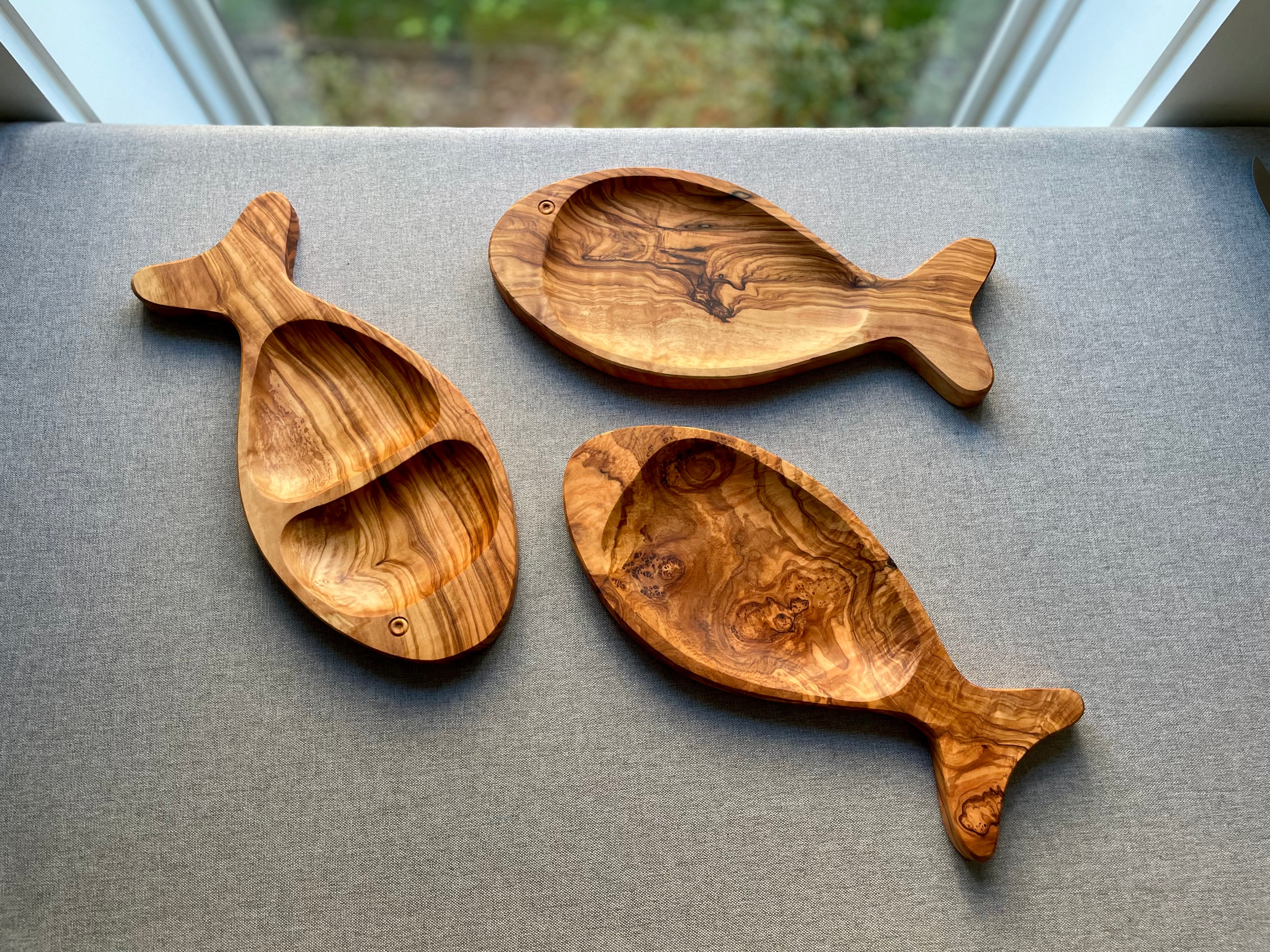 Three olive wood fish-shaped platters arranged on a bench by a window - hand-carved from natural olive wood, each with unique grain.